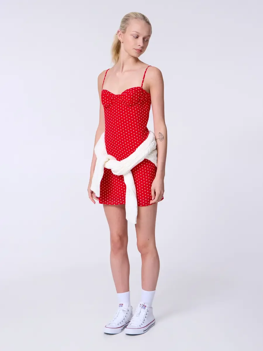 Model portfolio development session, commercial look, red polka dot sweetheart mini dress with cream chunky knit cardigan tied at waist, full-length, facing camera, gaze downward, cardigan knotted loosely at hip, sweetheart bodice bare above the knot