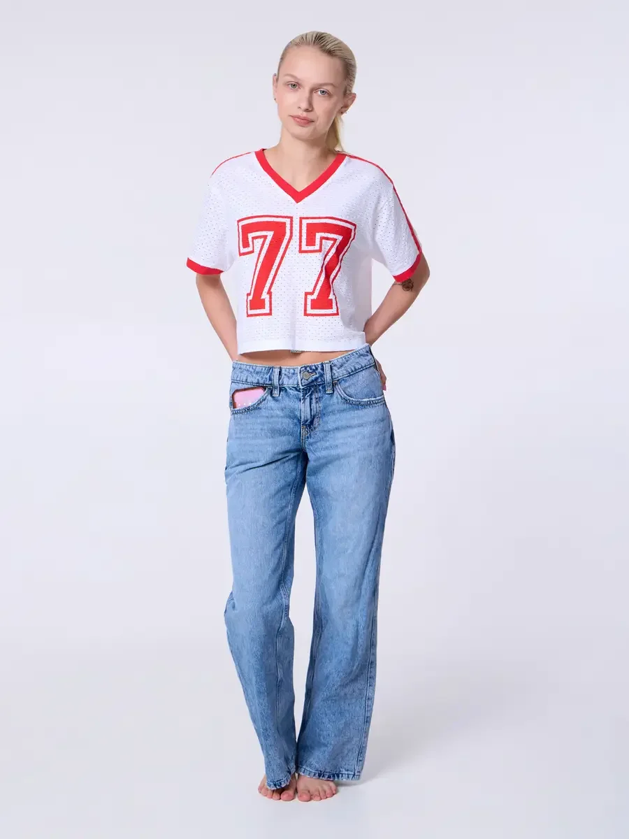 Model portfolio development session, commercial look, white mesh crop football jersey with red number 77 and red trim, low-rise light wash wide-leg jeans, full-length, facing camera, hands behind back, neutral expression, barefoot, light gray backgro