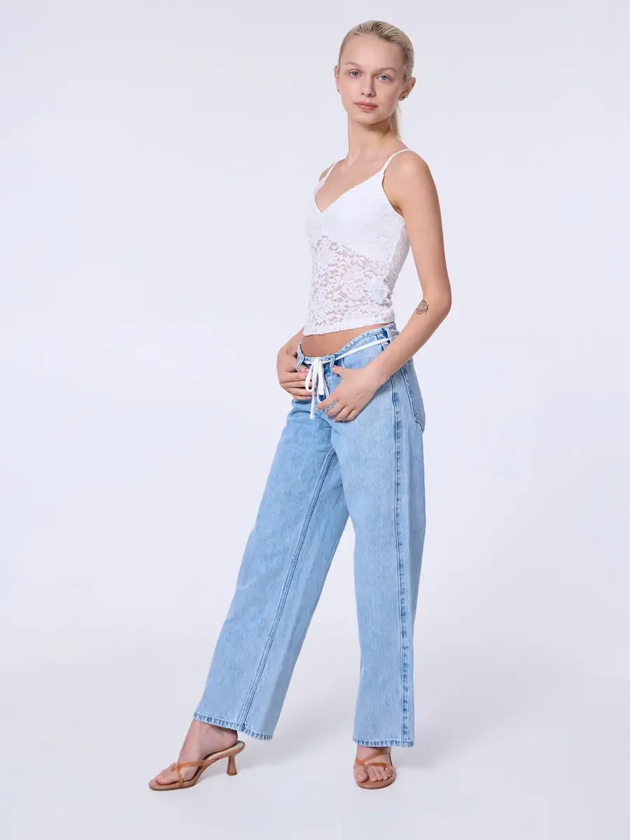 Model portfolio development session, commercial look, white lace crop camisole top and low-rise light wash wide-leg jeans with white drawstring detail, full-length, three-quarter turn to camera, one hand at waistband, relaxed stance, tan strappy heel
