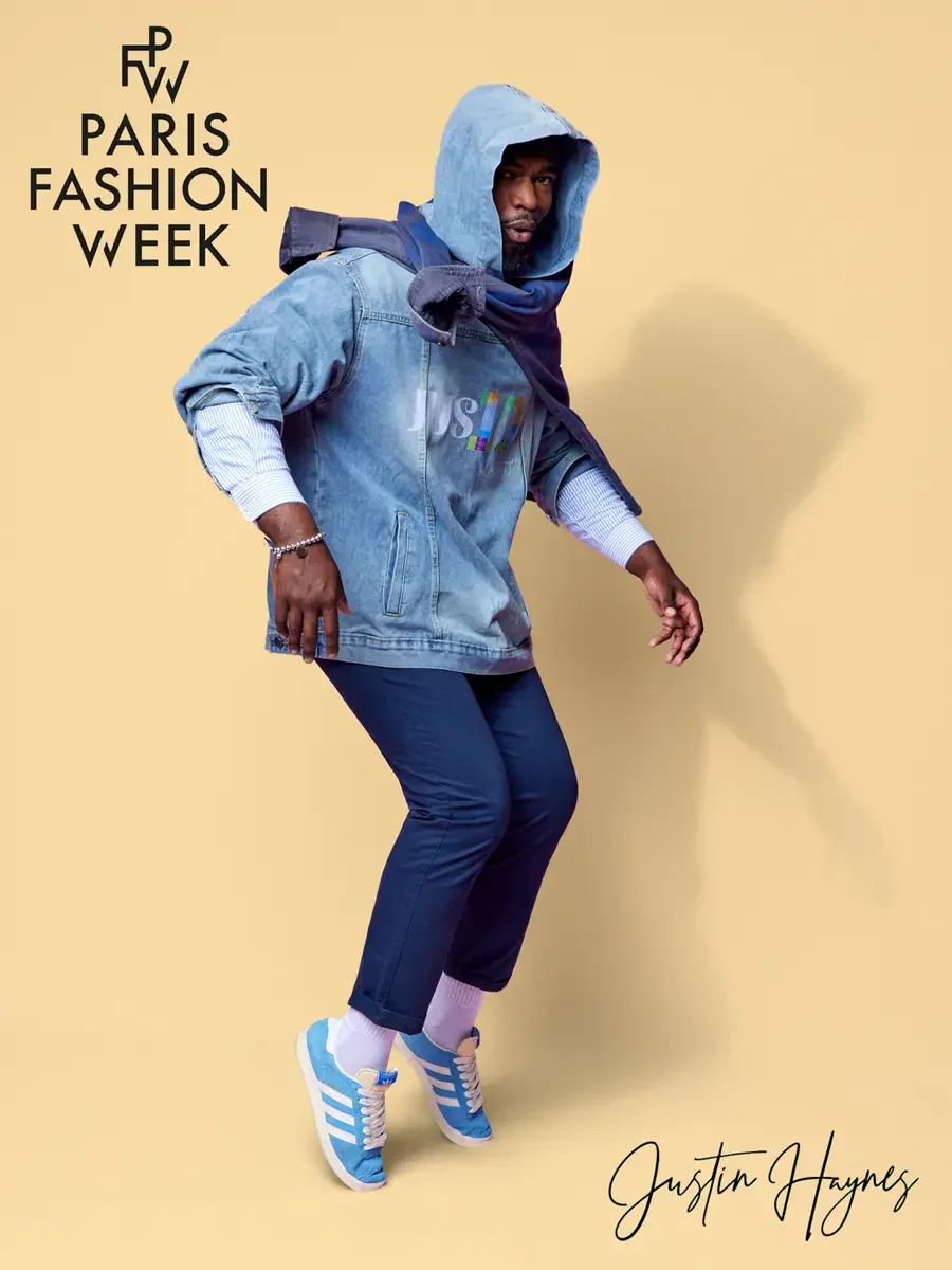 JUS10H 2025 promotional image, model mid-movement in light wash JUS10H logo denim jacket layered over navy hoodie and striped shirt, navy trousers, white socks and sky blue sneakers, butter yellow background, photographed by Scott Parker Photo