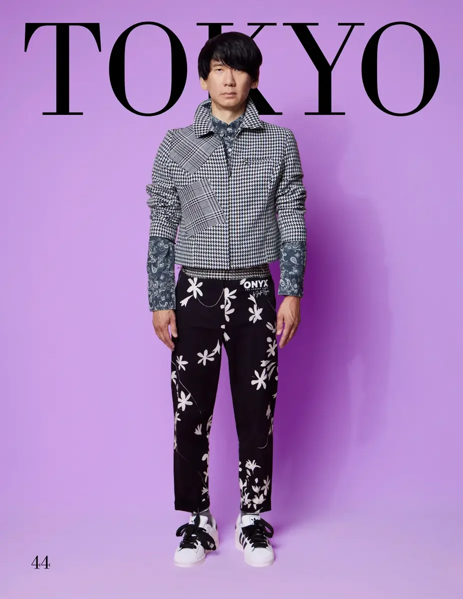JUS10H 2025 Tokyo lookbook, a male model wearing a black-and-white houndstooth and plaid patchwork zip-up jacket over a blue paisley shirt, paired with black trousers printed with a white floral pattern and an "ONYX" waistband logo, and white sneaker