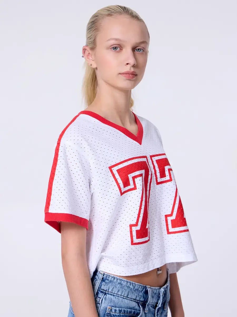Model portfolio development session, commercial look, white mesh crop football jersey with red number 77 and red trim, low-rise light wash wide-leg jeans, three-quarter, slight turn to camera, direct gaze, perforated mesh texture and red shoulder str