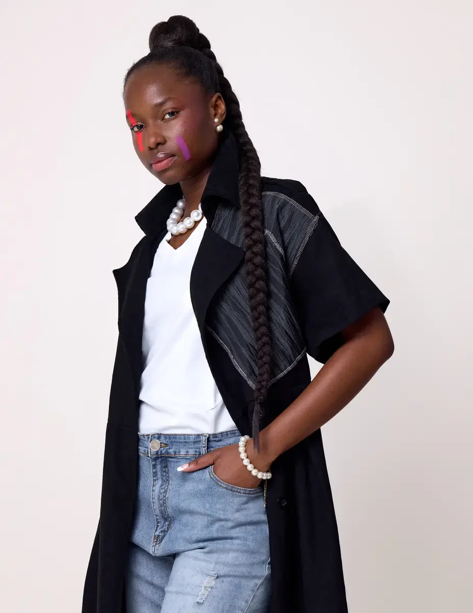 JUS10H Black Summer Jacket editorial, three-quarter portrait of female model in black long-line short-sleeve jacket with gray patchwork panels worn open, pearl necklace and bracelet, light-wash jeans, white background, photographed by Scott Parker Ph