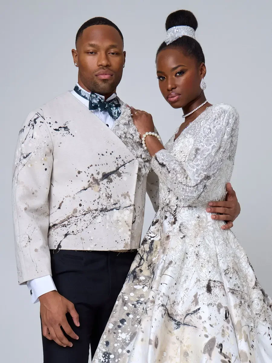 JUS10H 2025 New York Bridal Fashion Week, ivory abstract paint-splatter tuxedo jacket with dark teal floral bow tie and black trousers alongside white beaded lace ball gown with V-neckline, long sleeves, and embellished headband, styled with pearl ch
