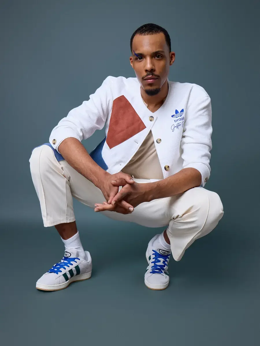  JUS10H x Adidas Brasil campaign, model in low squat with hands clasped, white textured patchwork cardigan with terracotta and blue suede panels over cream trousers, Adidas Campus 00s in white with dark green stripes and blue laces, steel blue backgr