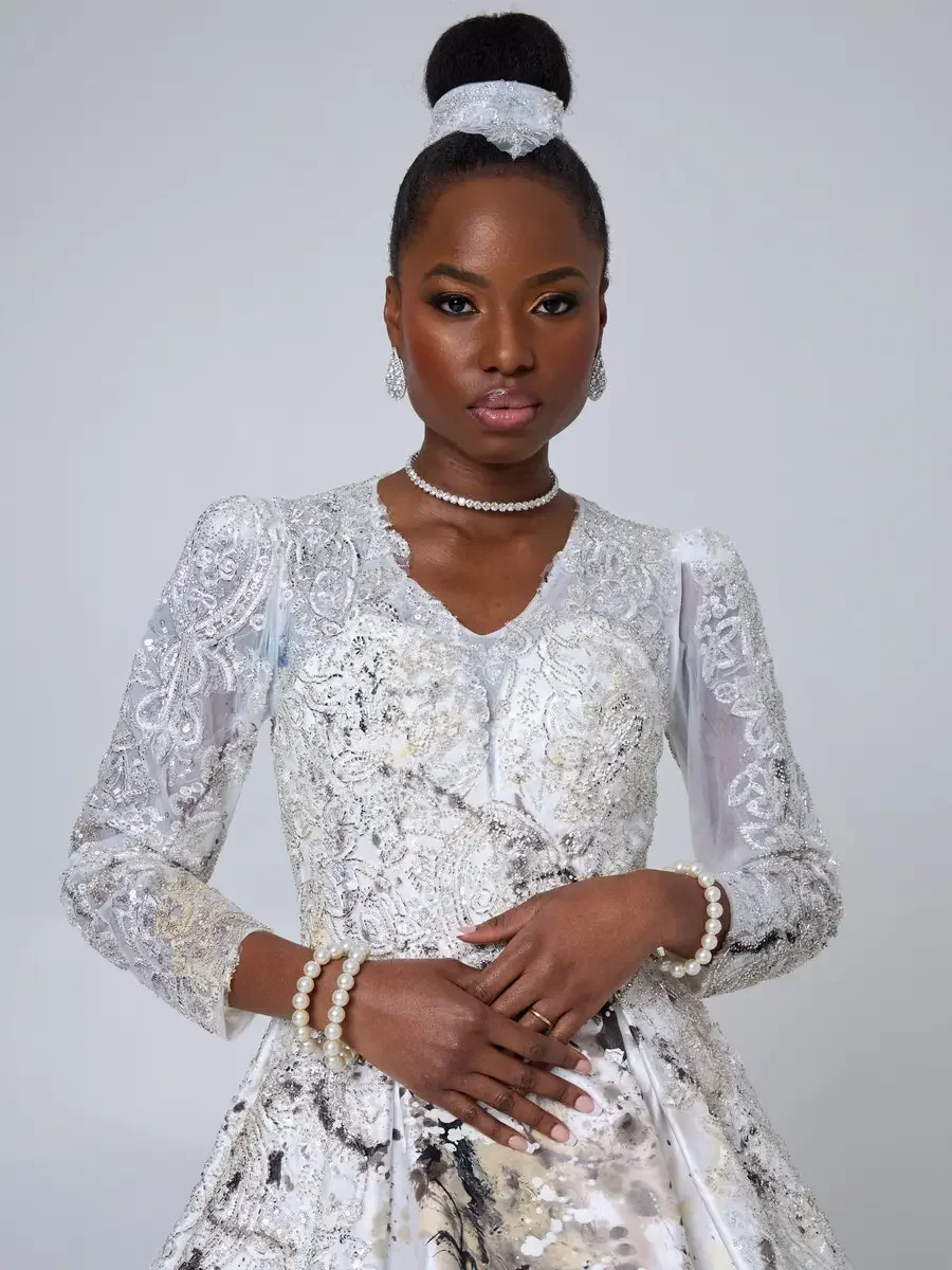JUS10H 2025 New York Bridal Fashion Week, detail of white silver-beaded lace ball gown with V-neckline, sheer embroidered puff sleeves, and embellished bun wrap, styled with diamond choker, drop earrings, and double pearl bracelet, photographed by Sc