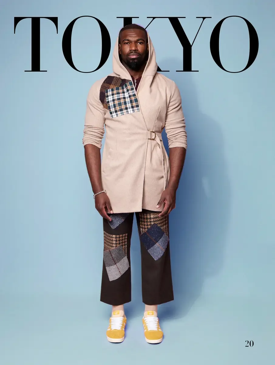 JUS10H 2025 Tokyo lookbook, cream belted hooded wrap cardigan with tartan patch over brown wide leg patchwork trousers with houndstooth and tweed panels and yellow Adidas sneakers, light blue background, photographed by Scott Parker Photo for the Tok