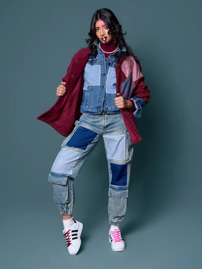 A woman with black curly hair and colorful makeup, wearing a layered denim jacket, multi-colored patchwork jeans, white sneakers with pink laces, and a maroon coat, standing against a gray background.