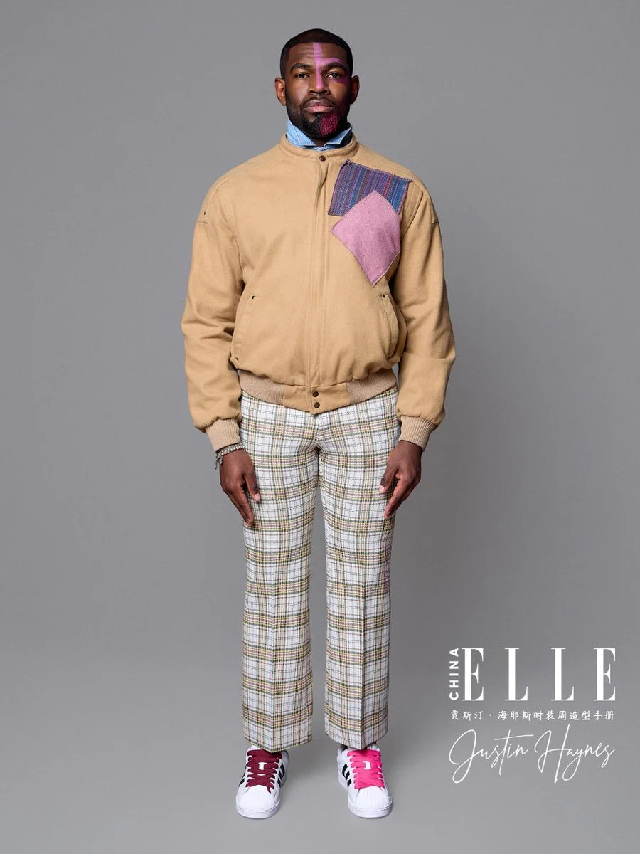 A man standing against a gray background wearing a beige jacket with colorful patches, plaid pants, and white sneakers with pink laces. He has a well-groomed beard and short hair, and is looking directly at the camera.