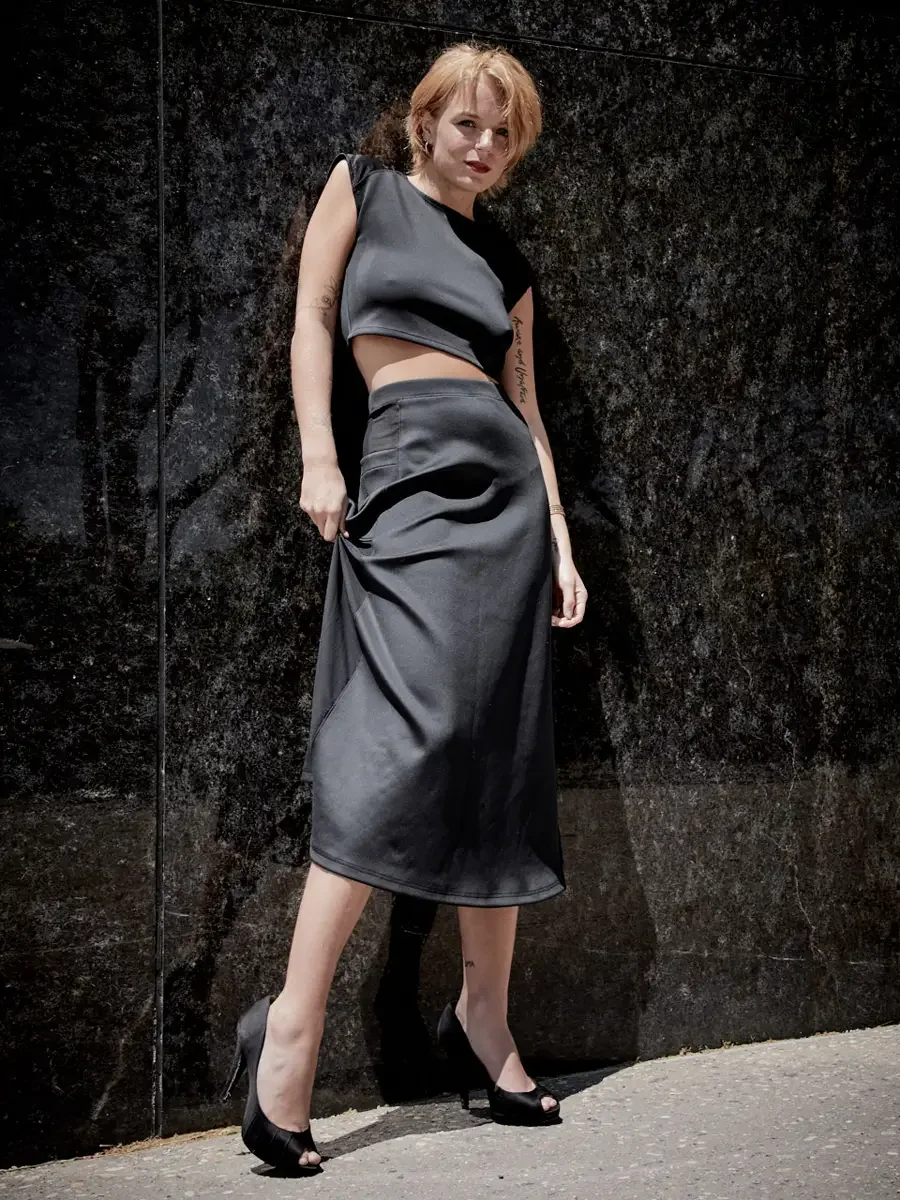 Satine, full length, one hand gathering skirt fabric, black structured cap-sleeve crop top, charcoal grey A-line midi skirt with sheer mesh hip panel, black satin peep-toe stiletto heels, script tattoo on right arm visible, granite wall, Battery Park