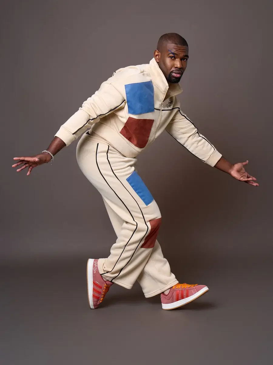 JUS10H x Adidas Brasil campaign, model in dynamic forward lean with arms extended, cream patchwork tracksuit with blue and terracotta suede panels and black piping, Adidas Campus 00s in pink suede with red stripes and orange laces, warm brown backgro