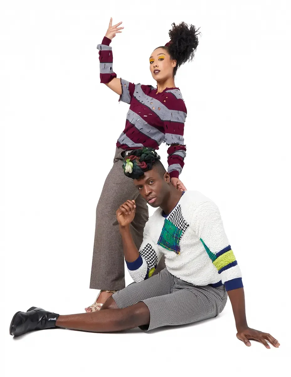 Model portfolio development session, high fashion editorial look, woman standing in burgundy and heather grey destroyed-knit stripe sweater and straight-leg heathered trousers with one arm raised, man seated on floor in white patchwork bouclé knit sw