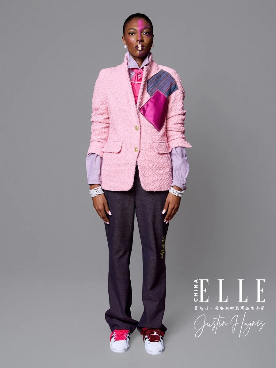A woman modeling fashion for a magazine cover, wearing a pink textured blazer over a lavender shirt, dark gray pants, and pink and white sneakers, with bold makeup and jewelry.