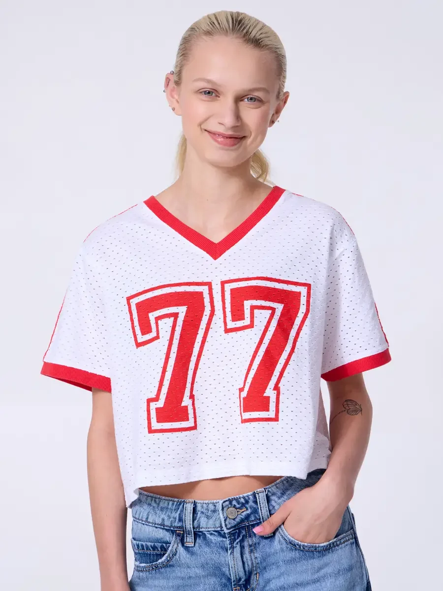 Model portfolio development session, commercial look, white mesh crop football jersey with red number 77 and red trim, low-rise light wash wide-leg jeans, three-quarter, facing camera, one hand in pocket, open smile, jersey number graphic and mesh te