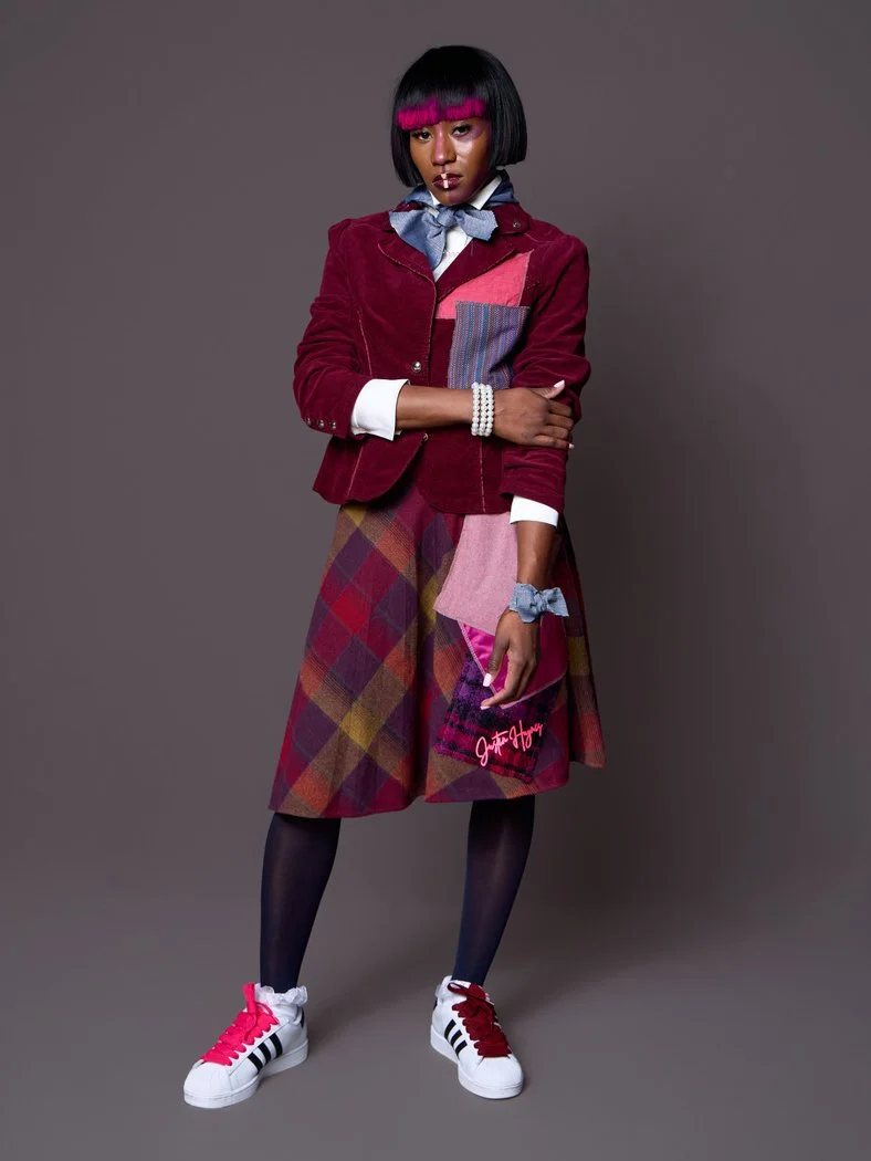 A woman with black hair and pink bangs wearing a red blazer, plaid skirt, black tights, and white sneakers with red laces, standing against a gray background.