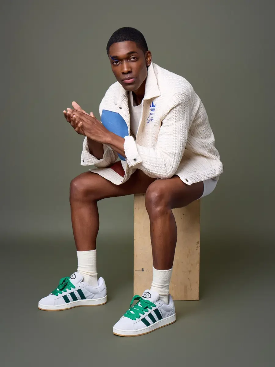 JUS10H x Adidas Brasil campaign, model seated on wood block with hands pressed together, white textured patchwork jacket with blue and terracotta suede panels, Adidas Campus 00s in white with dark green stripes and green laces, sage green background,