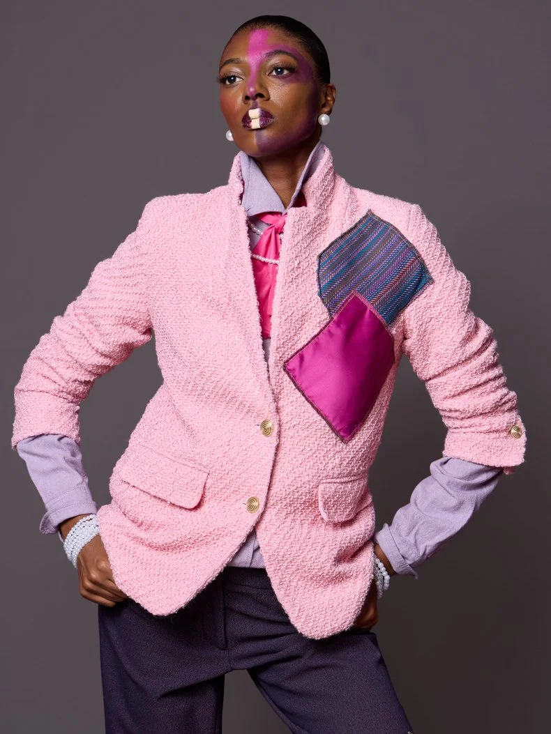 A woman with short hair wearing a pink textured blazer with a multicolored and pink pocket patch, lavender shirt with a pink necktie, dark pants, a pearl bracelet, and pearl earrings, standing against a gray background with a confident pose.