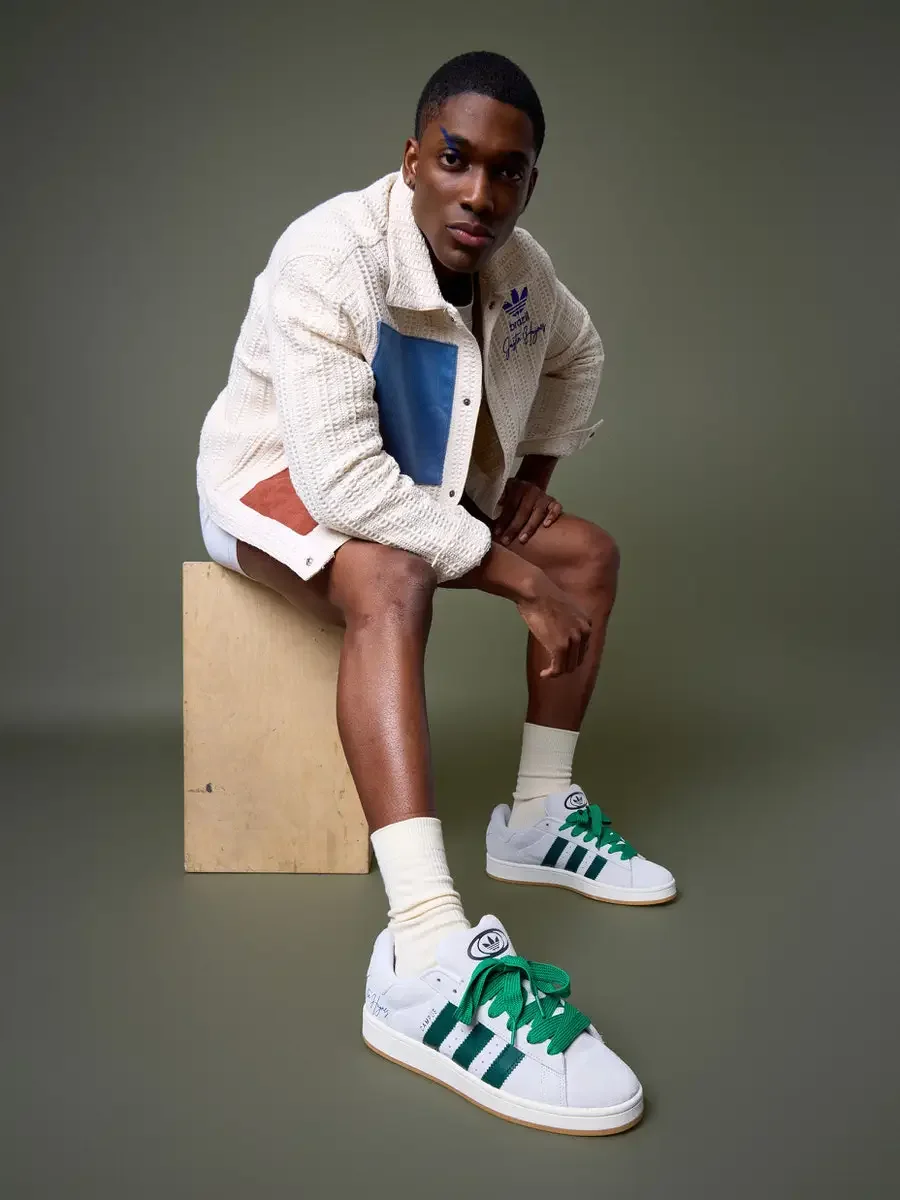JUS10H x Adidas Brasil campaign, model seated on wood block leaning forward, white textured patchwork jacket with blue and terracotta suede panels, Adidas Campus 00s in white with dark green stripes and green laces foregrounded, sage green background