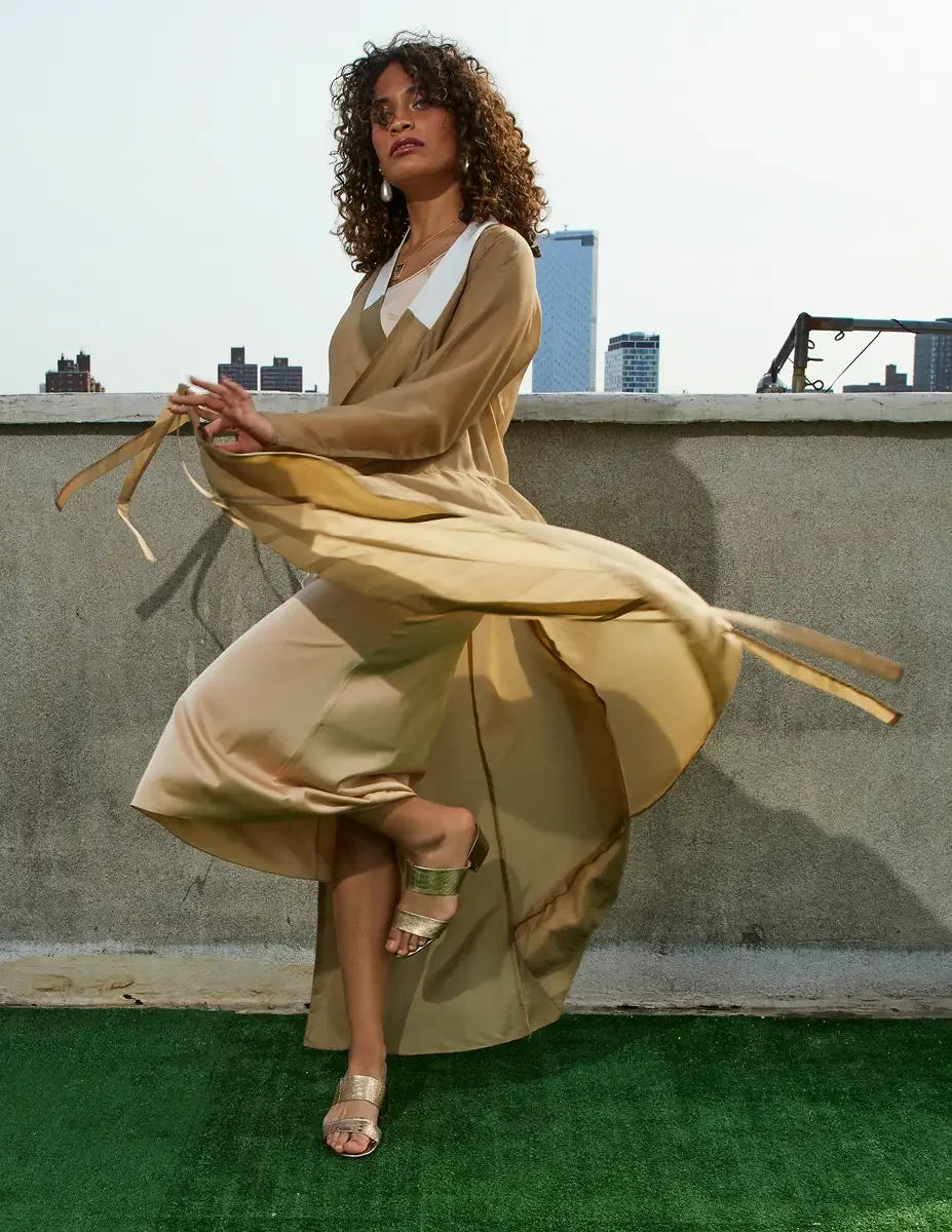 Fashion test shoot, full-length, model Ireland T in a gold satin wrap dress with tie belt in motion, Taro Collection earrings, Manhattan skyline, East Village rooftop, New York City, styling by Matsine Stinson, makeup by Stefani Paige Vivian, photogr