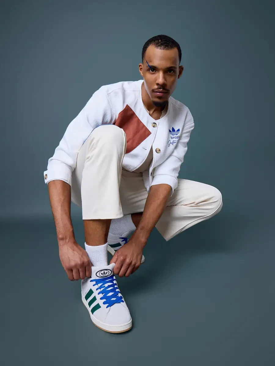 JUS10H x Adidas Brasil campaign, model lacing Adidas Campus 00s in white with dark green stripes and blue laces, white textured patchwork cardigan with terracotta suede panel over cream trousers, steel blue background, photographed by Scott Parker Ph