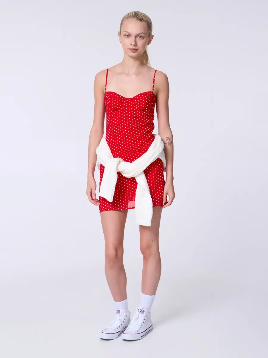 Model portfolio development session, commercial look, red polka dot sweetheart mini dress with cream chunky knit cardigan tied at waist, full-length, facing camera, arms relaxed at sides, direct gaze, cardigan knotted at hip showing full sweetheart b