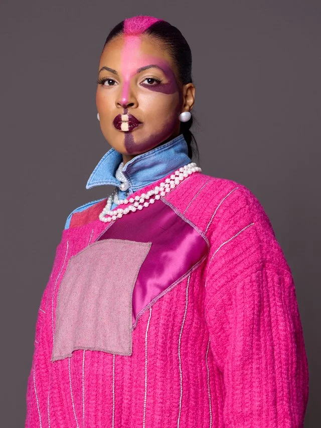 Woman with half her face painted pink and purple, wearing a pink textured jacket, pearl necklace, pearl earrings, and a denim collar underneath.