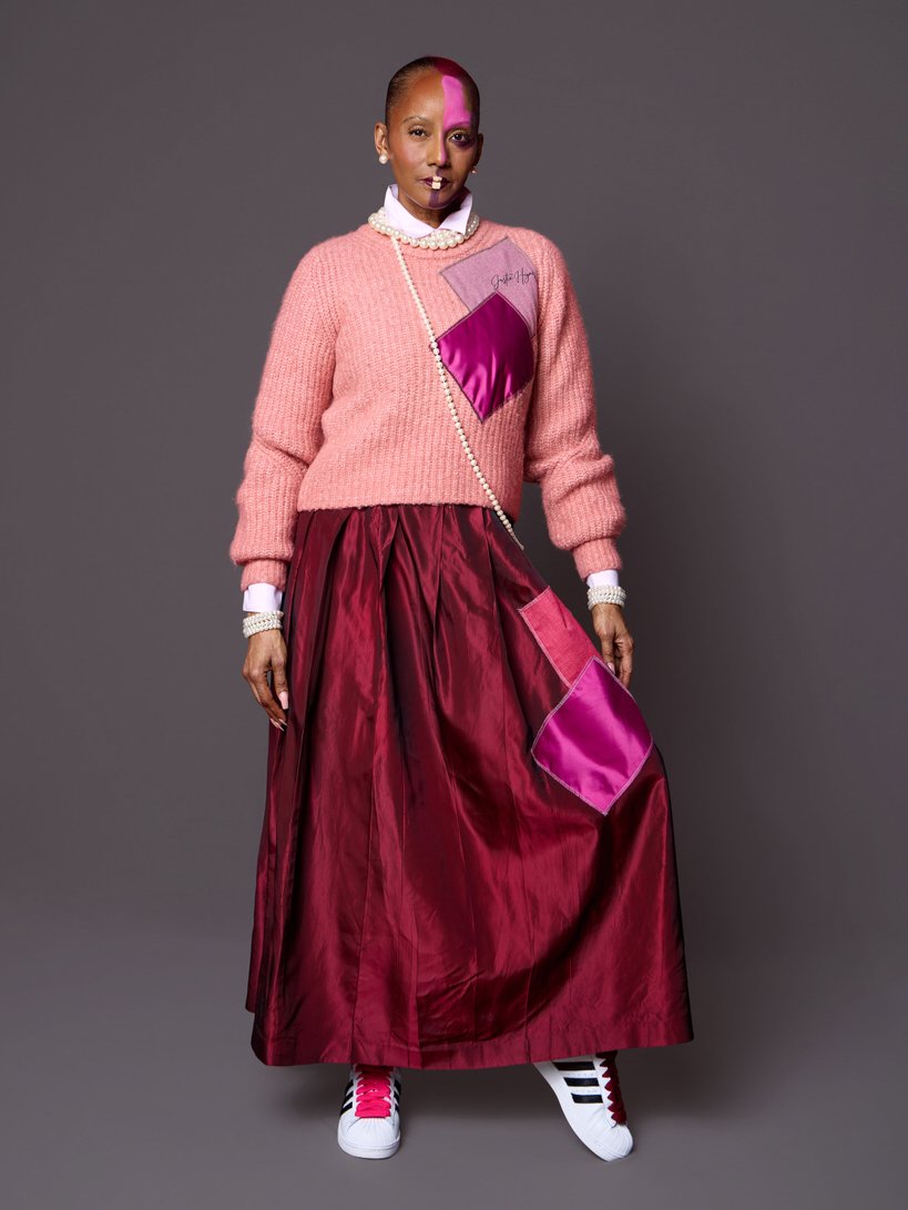 Woman wearing a pink sweater with patches, long maroon skirt, white sneakers with pink and black accents, pearl necklace, and earrings against a gray background.