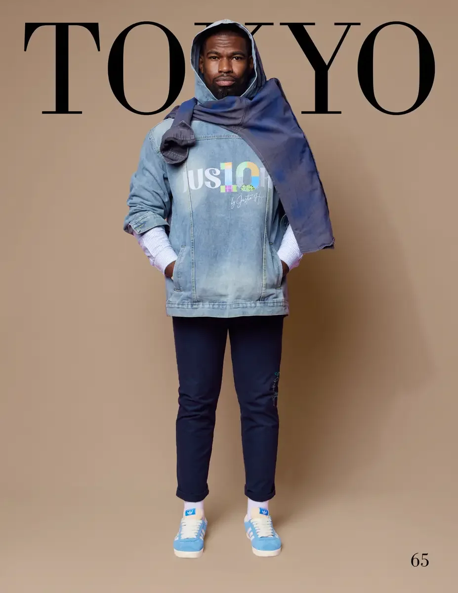 JUS10H 2025 Tokyo lookbook, a male model wearing an oversized light-wash denim jacket with a colorful holographic JUS10H logo graphic on the front, layered over a white and blue striped long-sleeve shirt, with a dark navy hooded sweatshirt draped ove