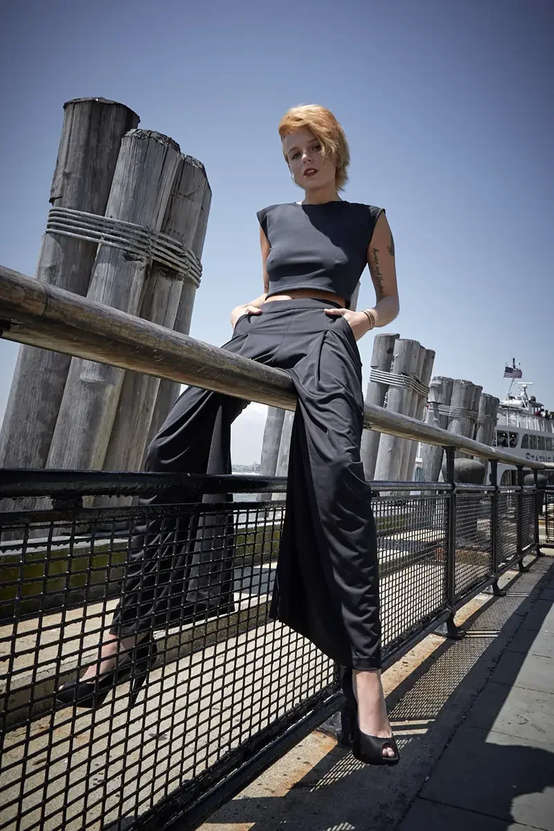 Satine, full length, standing, leg raised on pier railing, hands on waistband, black structured cap-sleeve crop top, black wide-leg pleated palazzo pants, black platform peep-toe heels, script tattoo on right arm visible, Battery Park pier test shoot