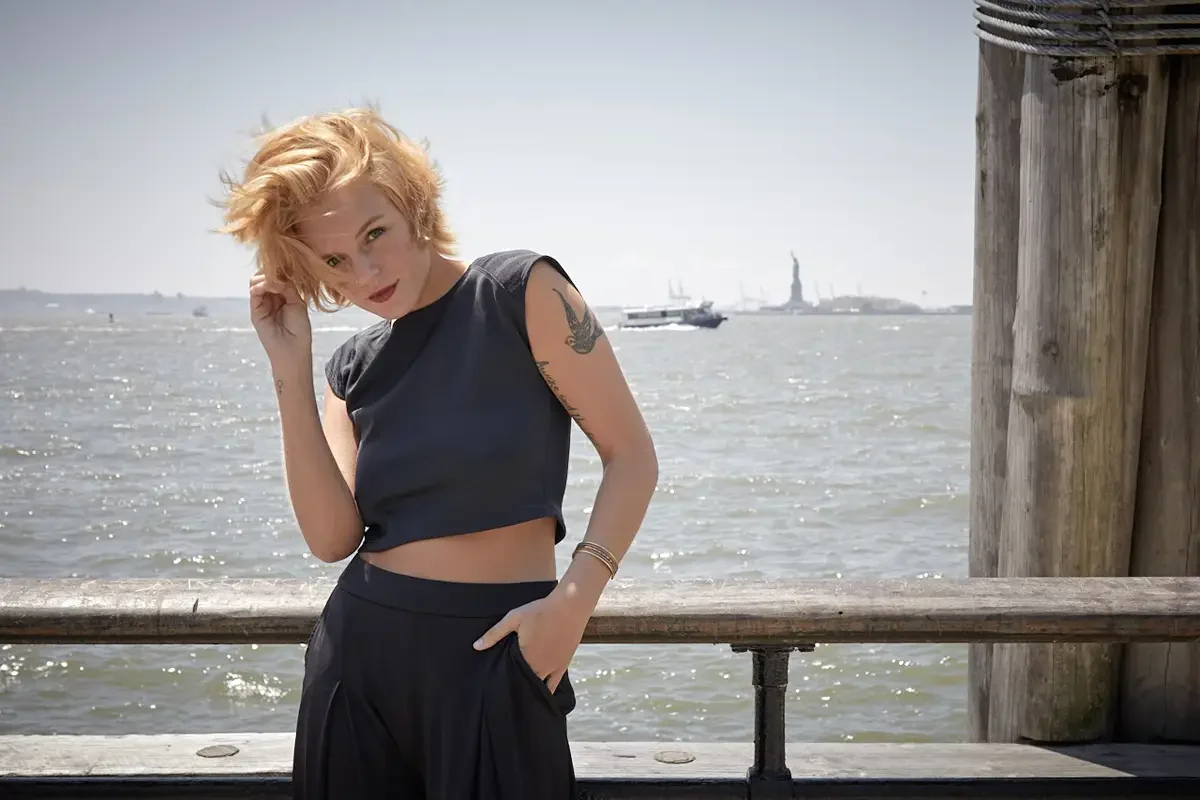 Satine, three-quarter crop, hand in hair, black structured cap-sleeve crop top, black wide-leg palazzo pants, gold bangles, bird tattoo on upper arm, Statue of Liberty visible in background, Battery Park waterfront test shoot, photographed by Scott P