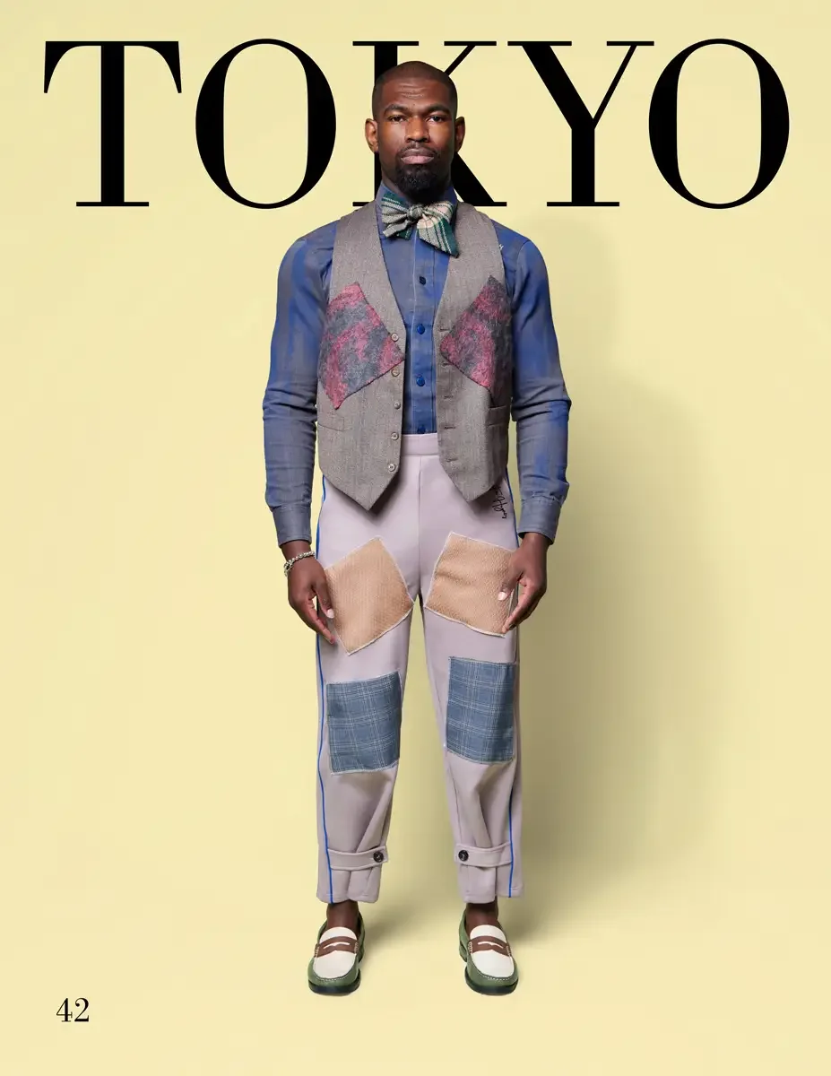 JUS10H 2025 Tokyo lookbook, a male model wearing a gray tweed vest with pink textured patchwork panels over a cobalt blue button-down shirt, lavender wide-leg trousers with tan and plaid patchwork appliqués and blue side stripes, a green plaid bow ti