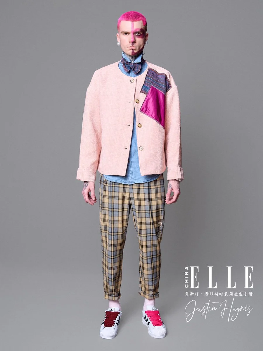 A male model with pink hair and face paint, wearing a pink jacket with colorful patches over a blue shirt, checkered beige pants, pink and white sneakers, and standing against a plain gray background, with ELLE China branding and Justin Hayes written