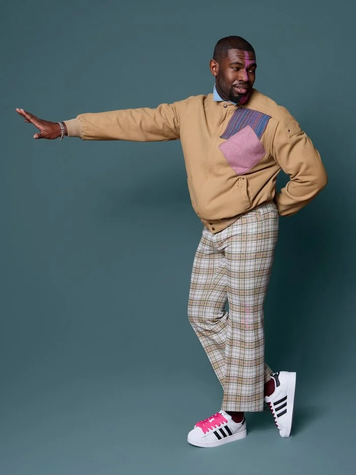 A man in a beige sweater with patches, plaid pants, and sneakers striking a playful pose against a plain teal background.
