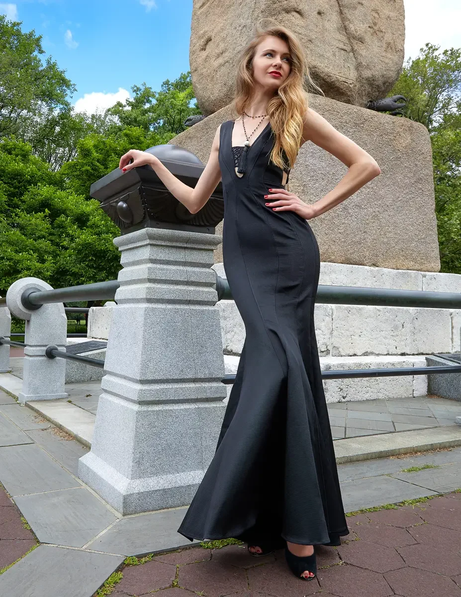 Julia, full length, standing beside a granite monument base with one arm resting on a bronze element, black bandage-style sleeveless mermaid gown with deep V-neckline, mesh lattice bodice detail, and floor-length flared hem, layered black bead choker