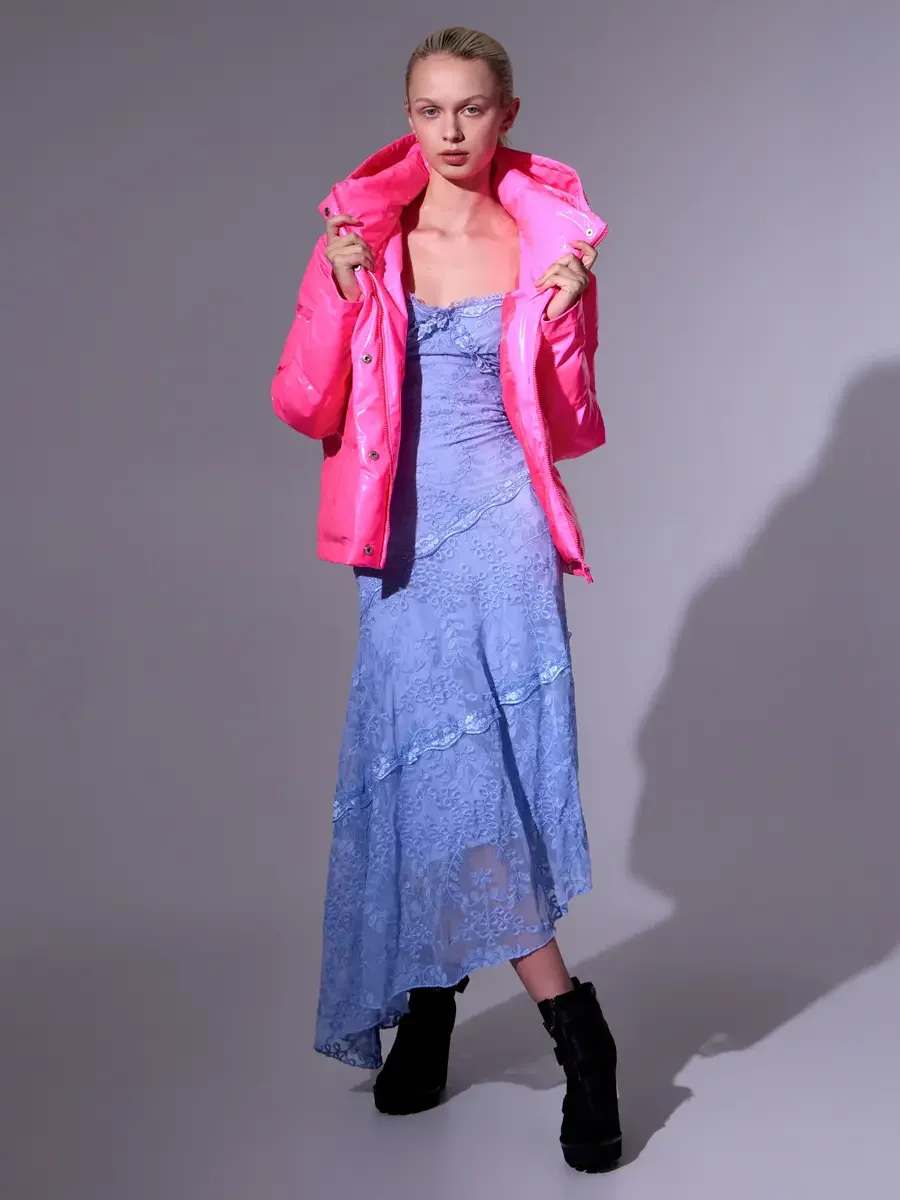 Model portfolio development session, high fashion editorial look, hot pink vinyl puffer jacket held open at collar over periwinkle blue lace asymmetrical midi dress, full-length, facing camera, both hands at lapels, composed expression, directional s