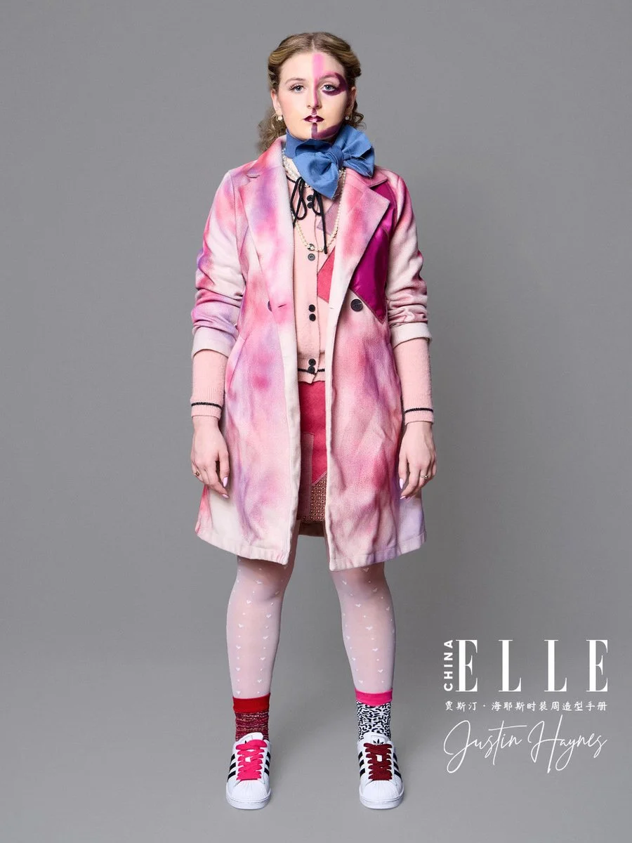 Young woman with pink and purple makeup, wearing a multicolored pink tie-dye blazer, pink skirt, patterned tights, sporty sneakers, and a large blue bow around her neck, standing against a plain grey background.