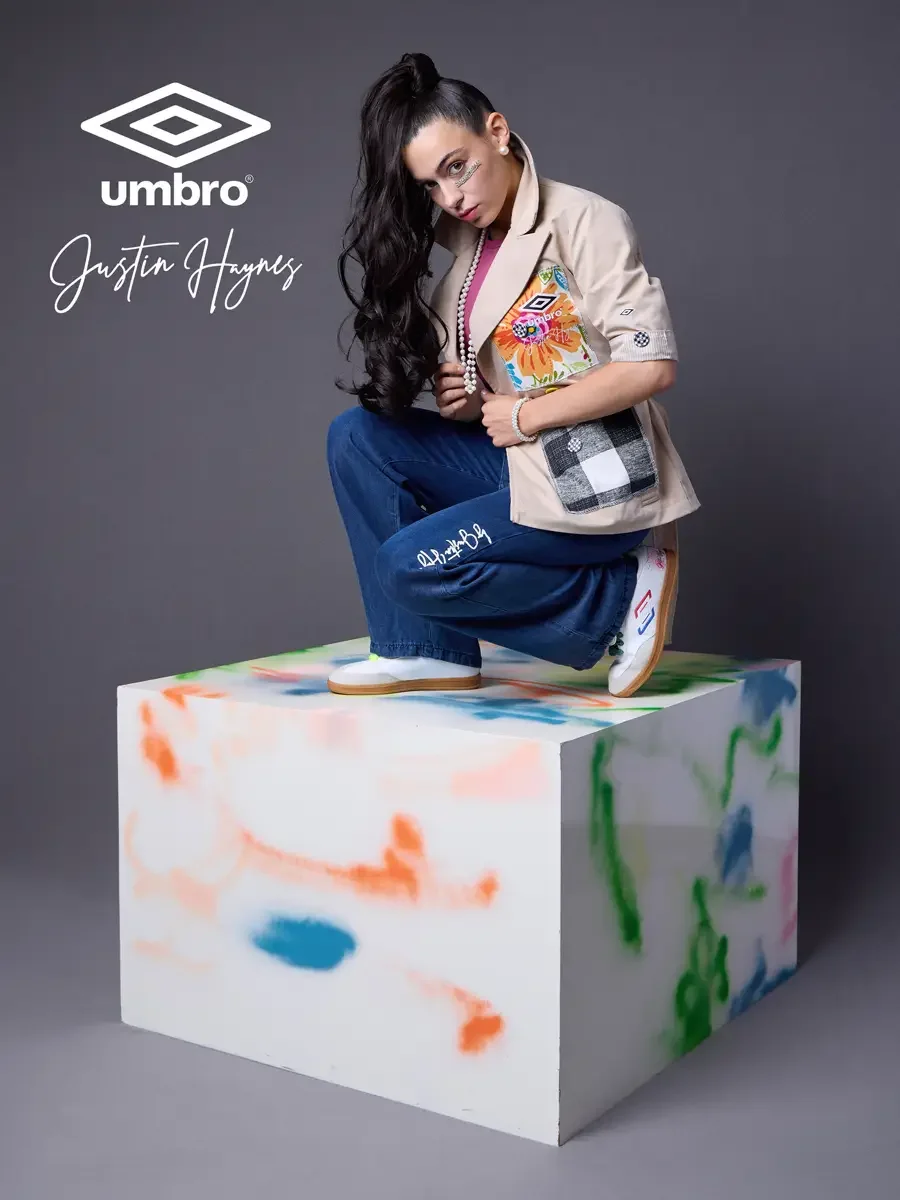 Polished pitch-side style for the Umbro France collection. Vanessa crouched low on a color-splashed prop in a sand utility jacket with floral patchwork panels over a fuchsia t-shirt and wide-leg dark indigo jeans, styled by Justin Haynes, photographe