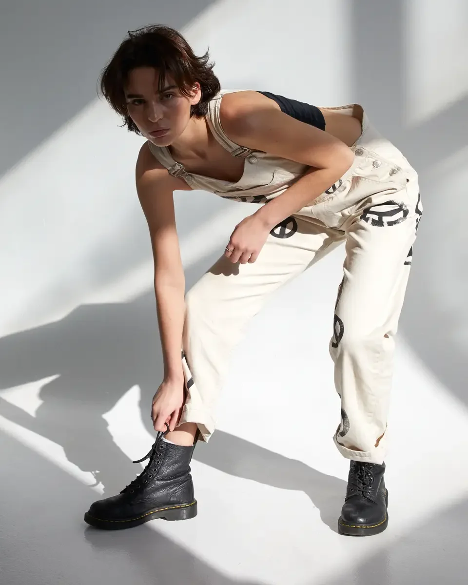 Tess, full-length, leaning forward with hands reaching toward boot, cream canvas overalls with allover hand-painted black peace sign print, black strapless bandeau top, black lace-up Dr. Martens boots, silver band ring, hard directional sunlight, New
