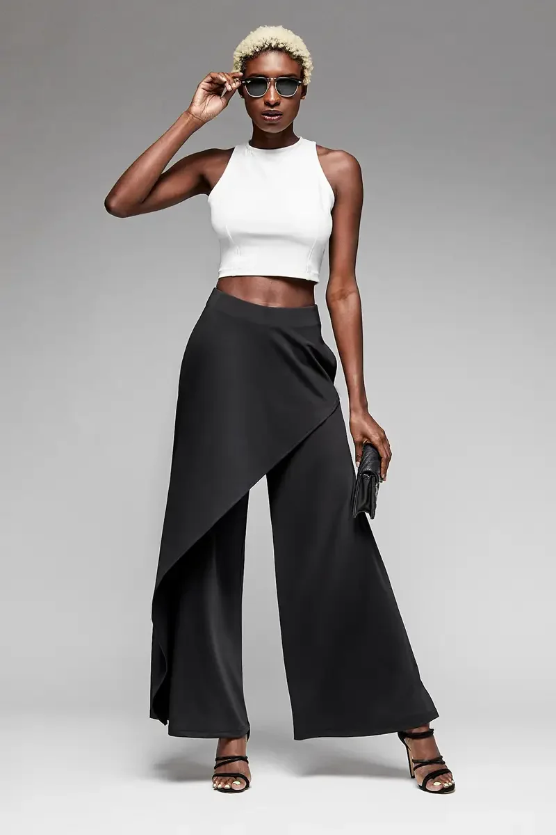Minika Ko lookbook, black wide-leg trousers with asymmetric wrap skirt overlay and white halter crop top, front view, high waist, draped skirt panel over wide-leg silhouette, smooth synthetic fabric, dark sunglasses, black clutch, photographed by Sco