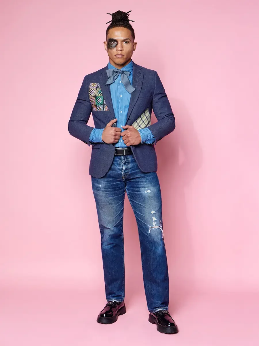JUS10H 2025 content library, model in navy blazer with multicolor embroidered and tartan patchwork panel over chambray shirt and denim bow tie, distressed indigo jeans and black patent lug-sole loafers, pink background, photographed by Scott Parker P