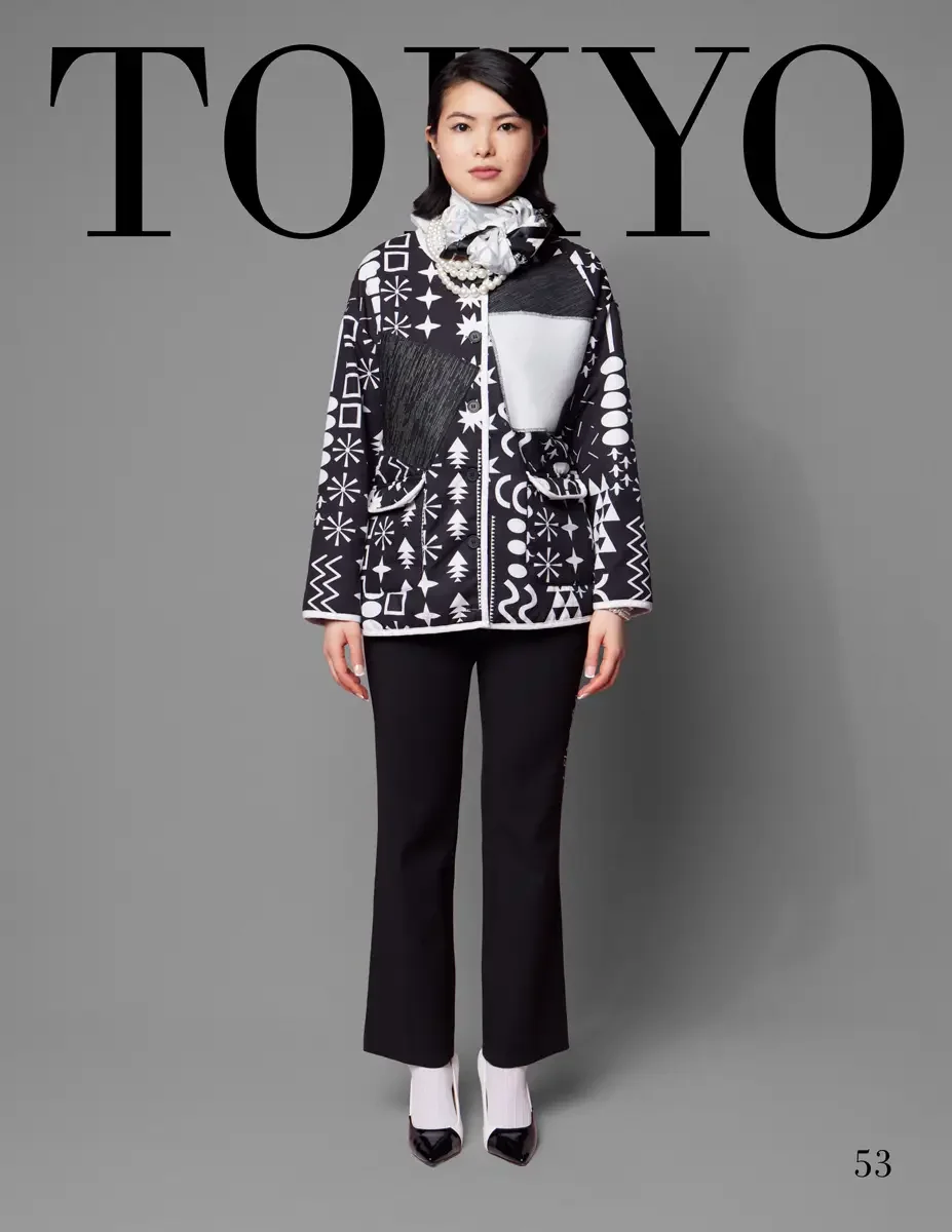 JUS10H 2025 Tokyo lookbook, a female model wearing a black and white graphic print jacket featuring stars, snowflakes, triangles, and geometric symbols, with black pleated and white color-block patchwork panels and a ruffled white satin scarf at the 