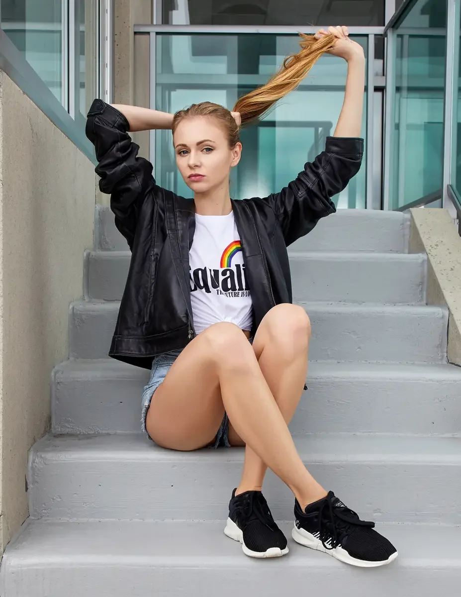 Julia, seated on rooftop access stairs with both arms raised holding hair aloft, white rainbow graphic tee, light-wash frayed denim cutoff shorts, oversized black zip-front leather jacket, black chunky sneakers with white soles, Jersey City rooftop t