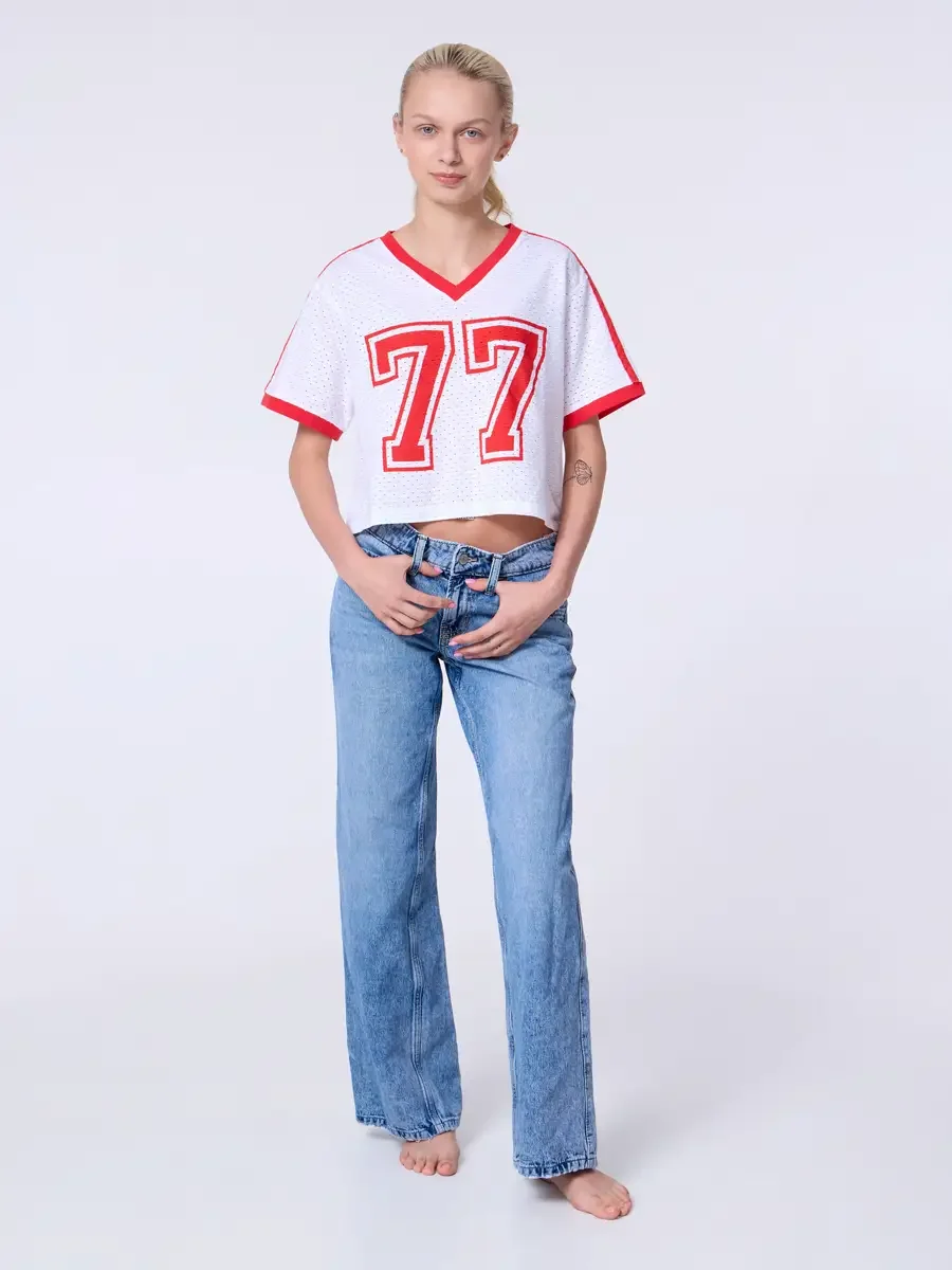 Model portfolio development session, commercial look, white mesh crop football jersey with red number 77 and red trim, low-rise light wash wide-leg jeans, full-length, facing camera, both hands at waistband, direct gaze, jersey graphic centered and f