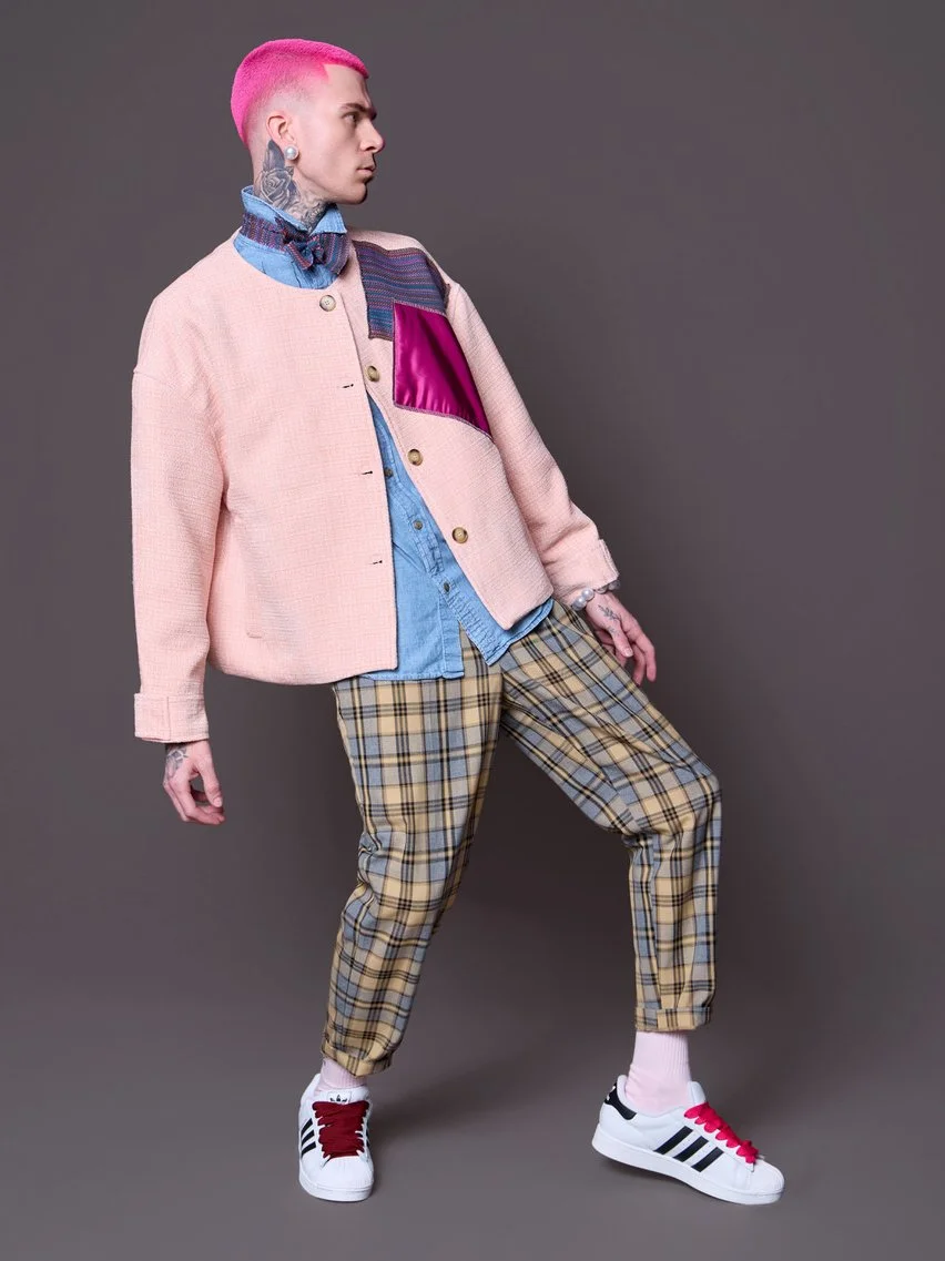 Person with short pink hair and tattoos wearing a pastel pink jacket, denim shirt, plaid pants, and sneakers, standing against a plain background.