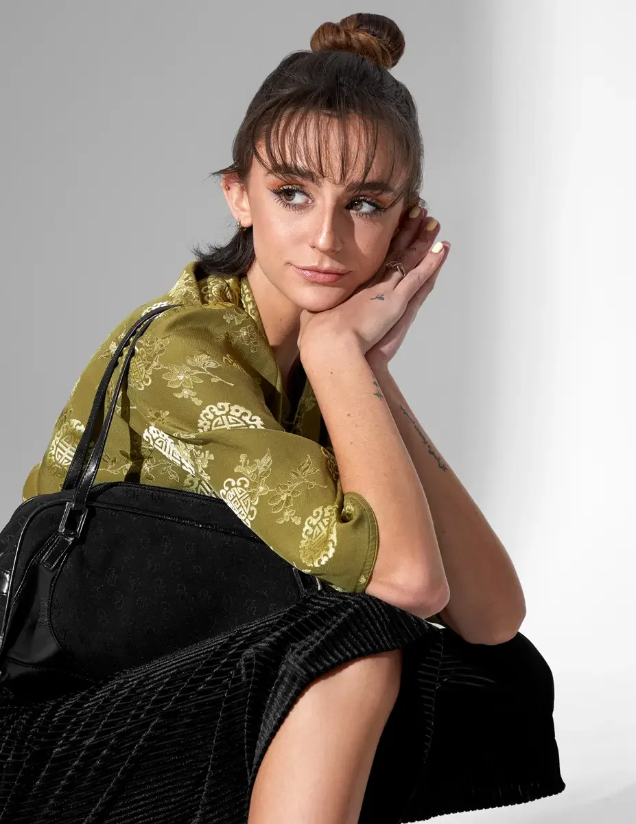 Macy, close crop, seated, hands together at chin, olive green satin jacquard crop top with gold medallion print, black wide-leg ribbed velvet culottes, black monogram barrel bag, script tattoo on inner forearm visible, New York studio test shoot, pho