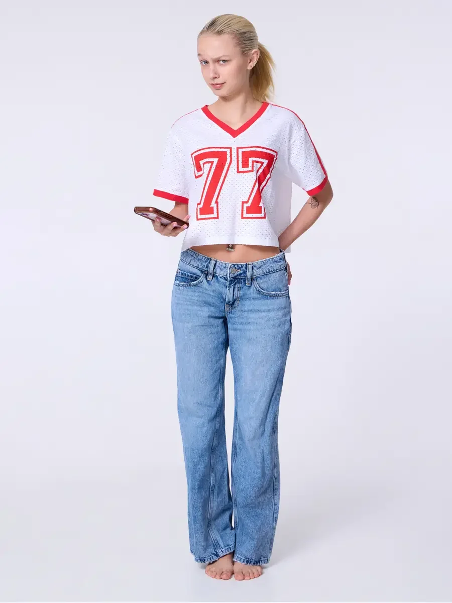 Model portfolio development session, commercial look, white mesh crop football jersey with red number 77 and red trim, low-rise light wash wide-leg jeans, full-length, slight turn, one hand holding phone at side, other hand in back pocket, composed e