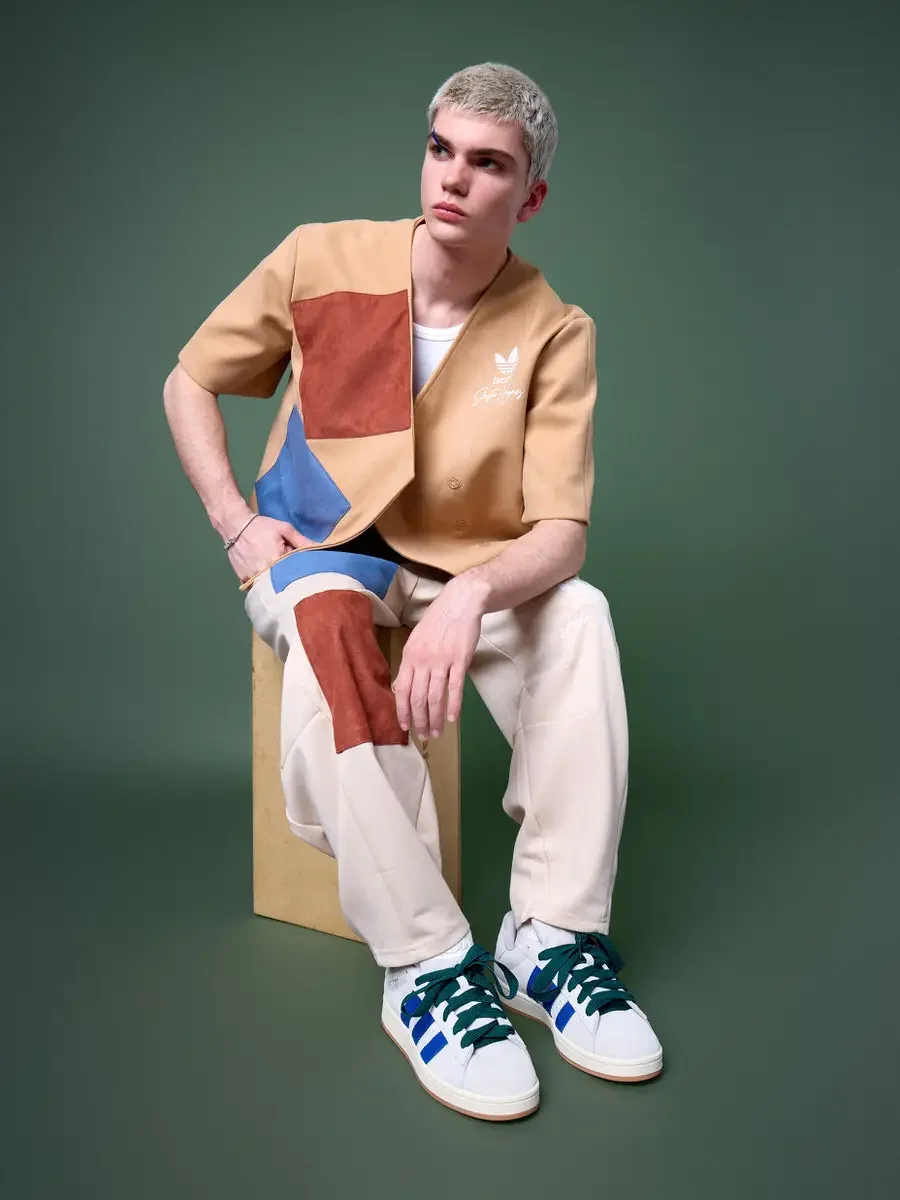JUS10H x Adidas Brasil campaign, model seated on wood block looking left, tan patchwork short-sleeve vest with terracotta and blue suede panels over cream wide-leg patchwork trousers, Adidas Campus 00s in white with blue stripes and dark green laces,