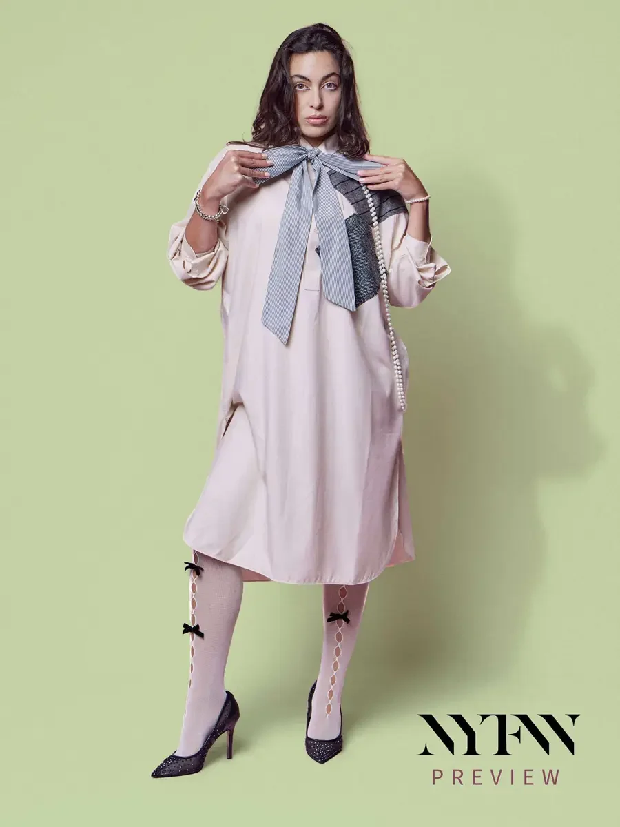 JUS10H 2025 print advertising, model in blush oversized shirt-dress with large striped bow and metallic patchwork panel, long pearl necklace, bow-detail sheer tights and glitter pumps, sage green background, photographed by Scott Parker Photo