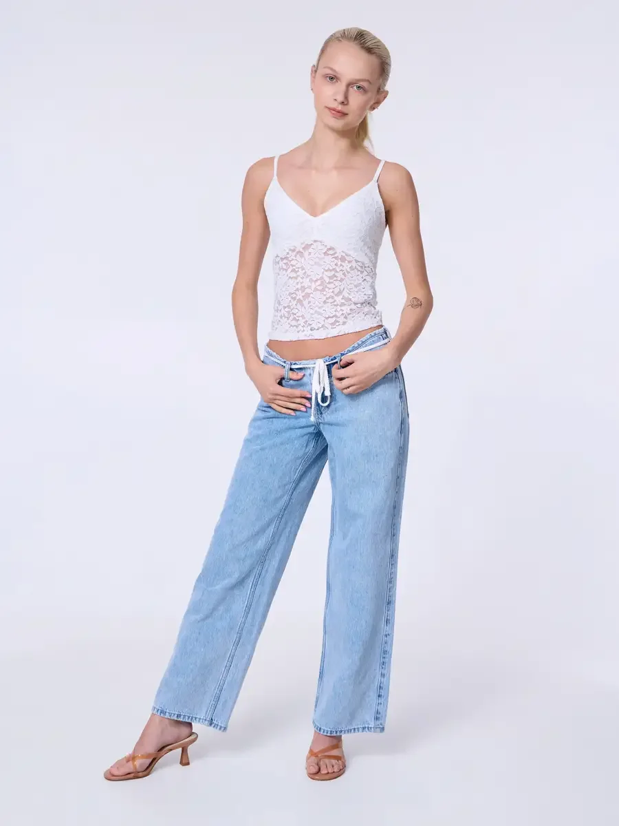 Model portfolio development session, commercial look, white lace crop camisole top and low-rise light wash wide-leg jeans with white drawstring detail, full-length, facing camera, hands tucked into front pockets, weight shifted to one hip, tan strapp