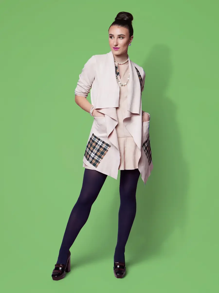 JUS10H 2025 Nylon Japan feature, model in blush waterfall coat with multicolor tartan patchwork pocket panel over blush drop-waist dress, layered pearl necklace, navy tights and burgundy platform loafers, green background, photographed by Scott Parke